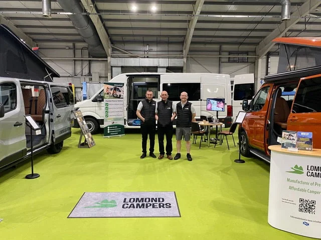 Three members from the Lomond Campers team standing in the middle of a large room, surrounded by their campervan conversion and motorhome conversion vans at a convention.